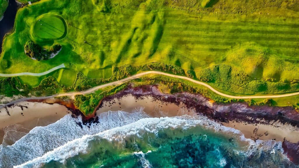 Stunning aerial view of a coastal golf course beside a vibrant beach with waves crashing onto the shore.