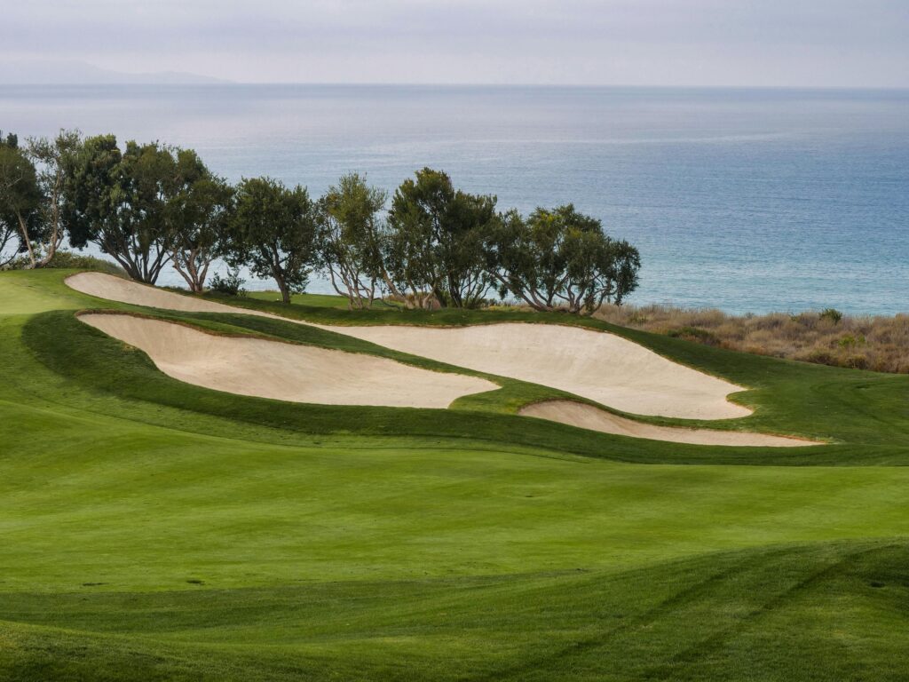 Golf course by the sea with green fairway and sandy bunkers under a cloudy sky.