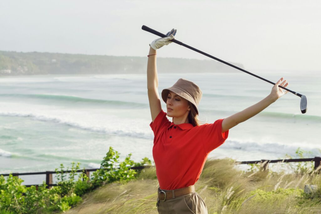 Woman golfer stretching with club by ocean, wearing red polo and hat.